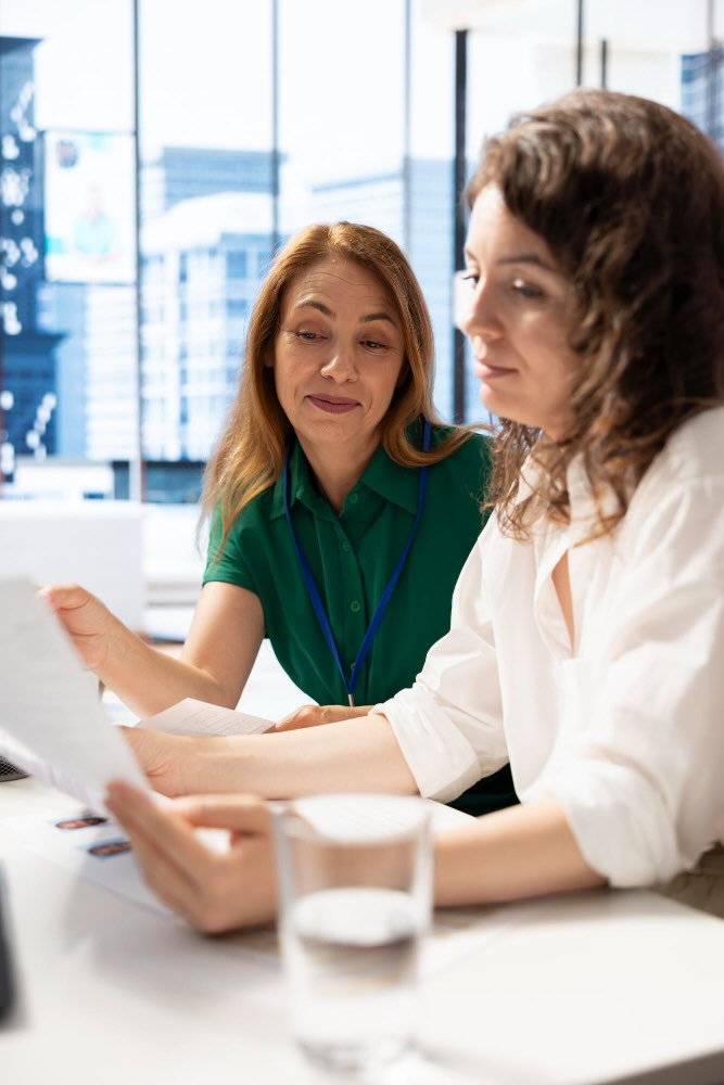Deux professionnelles en échange autour d'une table dans un bureau lumineux, examinant ensemble des documents / Témoignages sur l'accompagnement coaching santé et social avec IN FINE COACHING