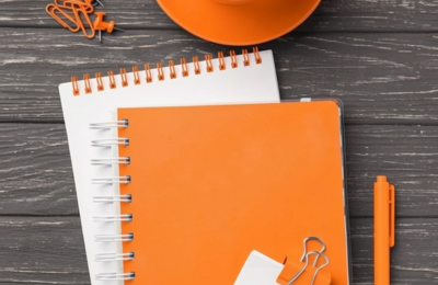 Carnets, stylo et trombones orange disposés sur une table en bois, prêts à préparer une session d'accompagnement - Les modalités d'intervention du coaching santé et social avec IN FINE COACHING